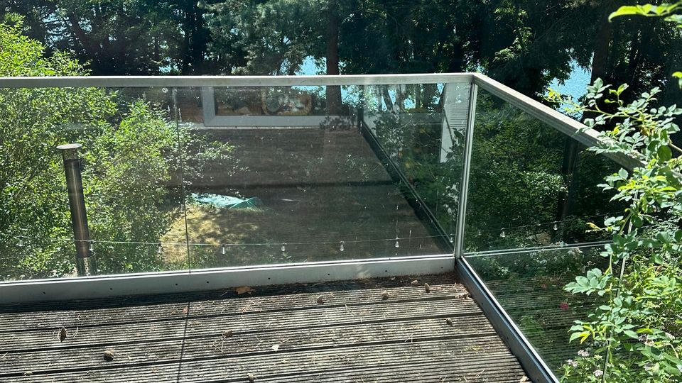 Holzterrasse mit geriffeltem Boden und transparenter Glasbrüstung mit Metallrahmen. Dahinter dichter Baumbestand mit Blick auf ein blaues Gewässer durch die Bäume.