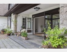 Eingangsbereich eines Hauses mit weißen Wänden, großen verglasten Türen und einem Vordach. Der gepflasterte Bereich ist mit Pflanzen in Töpfen und Körben dekoriert, darunter Blumen und Sträucher.