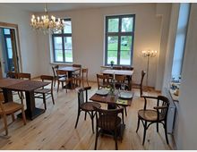 Heller Gastraum mit Holzboden und einfachen Holzmöbeln. Mehrere Tische und Stühle sind verteilt, ein Kronleuchter und ein Stehlicht sorgen für Beleuchtung, während zwei große Fenster den Blick nach draußen ermöglichen.