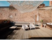 Eine Terrasse mit Holzboden vor einer Backsteinmauer. Auf der Terrasse stehen zwei weiße Stühle, ein niedriger Tisch, ein Sonnenschirm sowie Pflanzen in Töpfen, teils neben einer Sitzbank aus Rattan. Ein Sonnenschutznetz ist über der Terrasse gespannt.