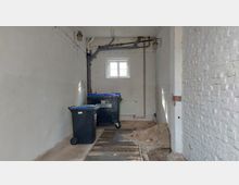 Ein schmaler, heller Raum mit weißen Backsteinwänden, einer kleinen Fensteröffnung und sichtbaren Rohren und Leitungen an der Decke. Im Vordergrund stehen Abfalltonnen, und der Boden zeigt Erdaushub und lose Backsteine.