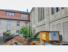 Außenansicht eines Gebäudekomplexes mit einer Fassade aus hellem Putz, großen vertikalen Fenstern und einem roten Ziegeldach. Im Vordergrund befinden sich Holzhackschnitzel, Metallstangen, ein gelber Maschinenkasten und etwas Grünbewuchs.