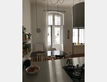 Küche mit hohen Decken und einem großen Fenster mit weißem Rahmen, das auf einen Balkon führt. Im Vordergrund befindet sich eine Kücheninsel mit Gaskochfeld und einer kleinen Espressokanne, dahinter ein Esstisch mit gemischten Stühlen.