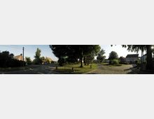 Ein ländlicher Dorfplatz mit einer gepflasterten Straße, einem Grünstreifen mit großen Bäumen und einem Wegweiser. Im Hintergrund sind Wohnhäuser mit roten Ziegeldächern und weißen Fassaden sowie vereinzelte Bäume sichtbar.