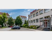 Straßenszene mit einem weißen Gebäude im rechten Vordergrund, auf dem ein rotes Schild mit der Aufschrift „Elektro“ angebracht ist. Im Hintergrund stehen Wohnhäuser mit roten Dächern und helle Fassaden, umgeben von Bäumen, sowie parkende Autos entlang der Straße.