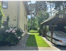 Seitlicher Blick auf ein helles Wohnhaus mit der Hausnummer 12, neben einem gepflasterten Weg und blühenden Sträuchern. Rechts steht ein hölzernes Carport mit überdachtem Stellplatz, dahinter ein grüner Garten mit hohen Bäumen.