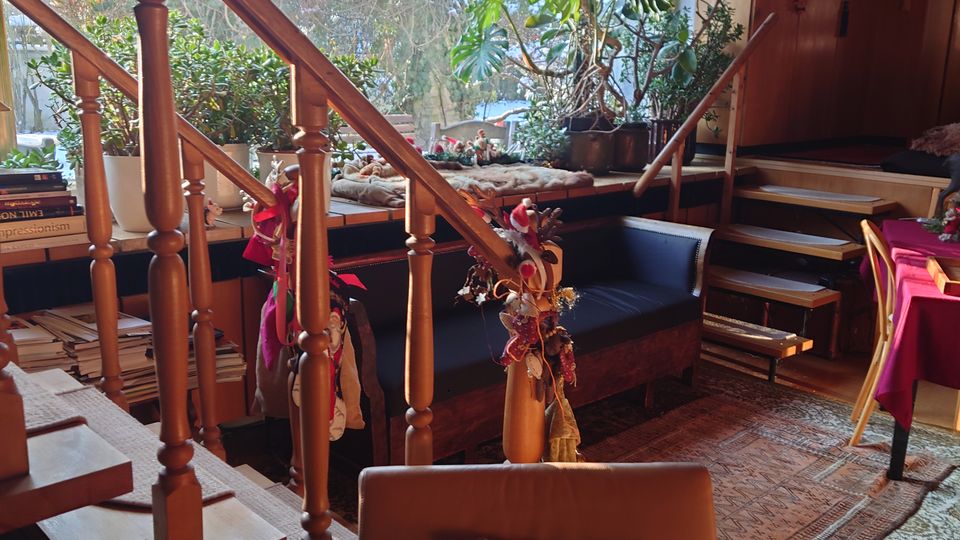 Wohnzimmer mit Holztreppe und großen Fenstern, die von Jalousien bedeckt sind. Im Raum befinden sich Pflanzen, gemütliche Sitzecken mit Kissen, Holzmöbel und ein Teppich auf Holzboden.