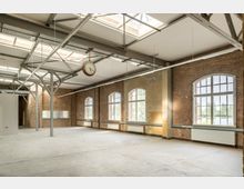 Großer, leerer Raum in einem ehemaligen Industriegebäude mit Backsteinwänden, hohen Decken und sichtbaren Stahlträgern. Große Fenster mit Rundbögen lassen viel Tageslicht hereinscheinen, während eine alte, hängende Uhr zentral im Raum zu sehen ist.