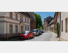 Gepflasterte Straße in einer Altstadt mit aufgereihten Autos und historischen Häusern auf beiden Seiten, darunter Fachwerk- und Backsteinbauten. Im Hintergrund sind Bäume und moderne Gebäude zu sehen.