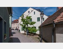 Schmale gepflasterte Gasse mit historischen Gebäuden, darunter ein helles Haus mit grünen Fensterläden und Ziegeldach. Ein Baum steht neben einer Straßenlaterne, und im Hintergrund sind weitere Häuser mit roten Dächern zu sehen.