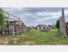 Freilichtmuseum mit einfachen Holzkonstruktionen und Nachbildungen von traditionellen strohgedeckten Hütten auf einer Wiese, umgeben von Bäumen unter einem bewölkten Himmel. Im Vordergrund sind unfertige Holzbauten und ein Zaun aus massiven Holzpfosten zu sehen.
