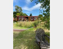 Ein rot gestrichenes, einstöckiges Gartenhaus mit weißen Fensterrahmen steht inmitten einer Wiese mit hohen Gräsern und Bäumen. Im Vordergrund sind eine Hängematte und ein Holzstuhl unter einem Baum zu sehen.