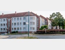 Mehrstöckige Wohngebäude mit hellgrauer Fassade, roten Ziegeln und roten Ziegeldächern, umgeben von einem runden Verkehrskreisel mit Gehwegen, Sträuchern und kleinen Bäumen. Im Hintergrund sind größere Bäume sichtbar, die entlang der Straße stehen.