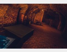 Gewölbekeller mit Backsteinwänden und -boden, spärlich beleuchtet durch warmes orangefarbenes Licht. Im Hintergrund stehen Holzbänke und Wände aus gestapelten Ziegeln, während ein moderner Tisch im Vordergrund sichtbar ist.