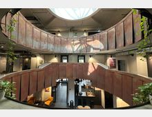 Offener Innenbereich eines modernen Gebäudes mit zwei Etagen, umgeben von rostbraunen Metallbrüstungen. Großes Oberlicht sorgt für natürliche Beleuchtung; Pflanzen und dekorative Möbel wie gelbe Sessel sowie eine offene Küche prägen das Design.