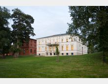 Eine historische Parklandschaft mit einem weißen, zweistöckigen Gebäude im klassizistischen Stil, das durch große Fenster und einen zentralen Balkon hervorsticht. Links daneben steht ein rotes Backsteingebäude im Industriestil, umgeben von großen Bäumen und einer Wiesenfläche.