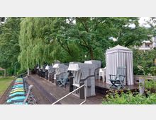 Ein Holzdeck mit mehreren Strandkörben und einer gestreiften Umkleidekabine, umgeben von großen grünen Bäumen. Im Vordergrund stehen bunte Sonnenliegen, und im Hintergrund sind Teile eines Gebäudes sichtbar.
