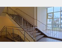 Innenansicht eines Treppenhauses mit zweiläufiger Treppe, grauen Stein- bzw. Betontreppen und weißen Metallgeländern mit Handlauf. Rechts sorgt ein großes, mehrteiliges Fenster für Tageslicht und bietet Blick auf umliegende Gebäude und Bäume.