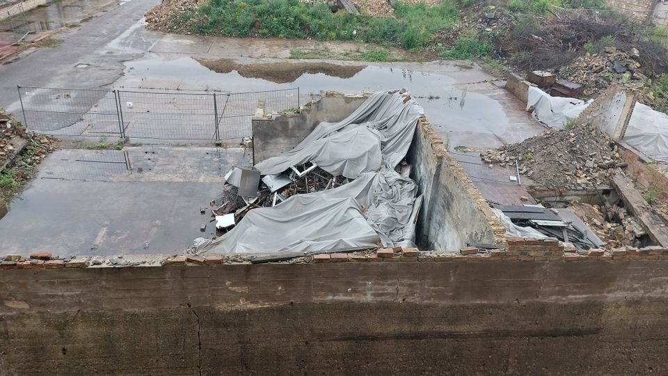 Ein verlassenes, teils eingestürztes Gebäude mit freiliegenden Mauern und Schutthaufen, bedeckt von grauen Planen. Der Boden ist stellenweise mit Wasser bedeckt, und das Gelände wirkt ungenutzt, umgeben von Vegetation und Trümmern.