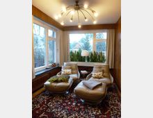 Wohnzimmer mit zwei beigefarbenen Ledersesseln und dazugehörigen Fußhockern auf einem gemusterten Teppich. Große Fenster mit hellen Vorhängen ermöglichen den Blick ins Freie, während Zimmerpflanzen die Fensterbank dekorieren; eine geschwungene Stehlampe sowie ein Kronleuchter mit mehreren Leuchten erhellen den Raum.