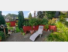 Ein gepflegter Garten mit einer Terrasse, auf der eine weiße Liege steht, umgeben von mehreren Kübelpflanzen, einem roten Mauersockel und hohen Sträuchern. Im Hintergrund sind ein Vogelhäuschen, Bäume und eine rote Gartenlaube sichtbar.