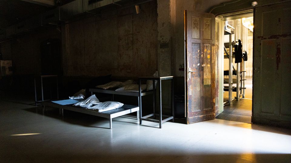 Dunkler, karger Innenraum mit rohen Betonwänden und sichtbaren Lüftungskanälen an der Decke, in dem zwei einfache Metallbetten mit Kissen stehen. Eine offene, abgenutzte Holztür führt in einen helleren Nebenraum mit industriellen Regalen und Geräten.