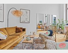 Wohnzimmer mit heller, moderner Einrichtung, bestehend aus einem senfgelben Sofa, Rattanstühlen mit bunten Kissen, einem Teppich und hellen Holzböden. Im Hintergrund ein großer Spiegel, der einen Essbereich mit Pflanzen, Tisch und Stühlen widerspiegelt.