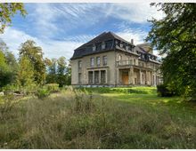 Eine alte, zweistöckige Villa mit abgenutzter Fassade und Mansardendach, umgeben von einer überwucherten Wiese und Bäumen. Das Gebäude wirkt leicht verfallen, mit sichtbaren Schäden an der Veranda und abgenutzten Fenstern.