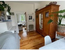 Heller Wohnraum mit Holzdielen, weißem Klavier an der linken Wand und großem Holzkleiderschrank mit Spiegeltür rechts. Eine offene Glastür führt in ein angrenzendes Wohnzimmer mit Sofa und Fenster; an den Wänden hängen Fotos und Bilder, dazu stehen Zimmerpflanzen.