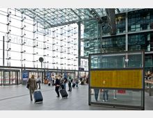 Großes, modernes Bahnhofsgebäude mit einer gläsernen Fassade und Stahlträgern, durch die viel Tageslicht einfällt. Menschen mit Gepäck laufen durch die Halle, im Vordergrund eine Tafelkonstruktion mit gelben Zugfahrplänen.