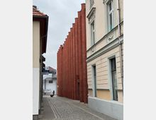 Schmale Gasse mit Kopfsteinpflaster, flankiert von einem modernen roten Ziegelgebäude mit abgestuften Fassadenelementen auf der linken Seite und einem älteren, verputzten Wohngebäude mit Stuckdetails auf der rechten Seite. Im Hintergrund ist ein weißes Gebäude und ein Passant zu sehen.