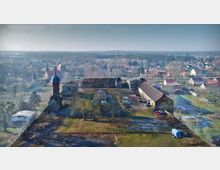 Luftaufnahme eines dörflichen Areals mit einem historischen Kirchen- oder Turmgebäude mit dunkler Kuppel, umgeben von Wiesen, Bäumen und mehreren länglichen Wirtschaftsgebäuden. Im Hintergrund erstreckt sich ein Wohngebiet mit Einfamilienhäusern und Feldern bis zum Horizont.