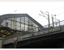 Außenansicht des Bahnhofs Friedrichstraße in Berlin mit einer gläsernen Stahlkonstruktion im oberen Teil und Schienenleitungen im Vordergrund. Ein Schild mit der Aufschrift "Bahnhof Friedrichstraße" und das Logo der Deutschen Bahn sind sichtbar.