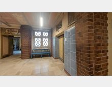 Ein Flur in einem historischen Gebäude mit Wänden aus rotem Backstein, einer Fensterreihe mit rautenförmigen Gittern und blau gefliesten Wandbereichen um eine Fahrstuhltür. Eine Holzbank steht unter den Fenstern, und oberhalb des Eingangs links ist ein Schild mit der Aufschrift "Kleine Schwimmhalle" angebracht.