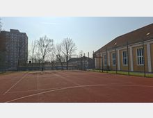 Eingezäunter Sportplatz mit rotem Kunststoffbelag und aufgemalten Spielfeldlinien, umgeben von kahlen Bäumen. Rechts steht ein langes, gelbes Gebäude mit Satteldach und hohen Fenstern, links ist ein mehrstöckiges Hochhaus im Hintergrund zu sehen.