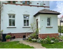 Fassade eines weißen Wohnhauses mit roten Backsteinelementen und einem kleinen, überdachten Eingangsbereich. Vor dem Haus befindet sich ein gepflegter Garten mit Blumenbeeten, einer Terrasse, und Gartenmöbeln.