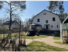 Großer Garten mit gepflastertem Weg, umgeben von Rasenflächen und Bäumen. Im Hintergrund steht ein großes, weißes Wohnhaus mit Satteldach, einer Holzterrasse und einem angrenzenden Gewächshaus rechts.