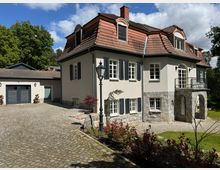Zweistöckiges Wohnhaus im klassischen Stil mit hellem Putz, einem roten Mansarddach und schwarzen Fensterläden, davor ein gepflasterter Hof und ein Laternenpfahl. Auf der linken Seite befindet sich eine angeschlossene Garage, im Hintergrund sind Bäume und ein heller Himmel sichtbar.