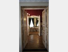 Blick durch geöffnete weiße Doppeltüren in einen Raum mit einem großen Esstisch, der mit weißen Hussenstühlen umgeben ist. Der Raum hat rote Wände, dunkle Vorhänge und hölzernen Fischgrätboden.