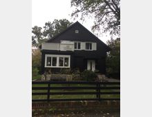 Zweistöckiges, schwarzes Haus mit weiß gestrichenen Fensterläden und einem Balkon im oberen Stockwerk. Das Gebäude steht in einem umzäunten Garten mit Rasenfläche, Büschen und Bäumen im Hintergrund.