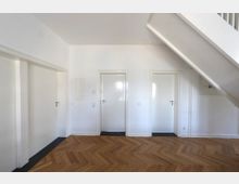 Ein heller Flur mit weiß gestrichenen Wänden, drei weißen Türen mit Türgriffen, einer hölzernen Chevron-Parkettboden, und einer weißen Treppe, die nach oben führt.