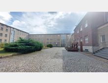 Weitläufiger Hof mit gepflastertem Boden, umgeben von mehrstöckigen Gebäuden aus rotem Ziegel und beigem Putz mit zahlreichen Fenstern; im Vordergrund Büsche und eine Metallinstallation, rechts eine Treppe.