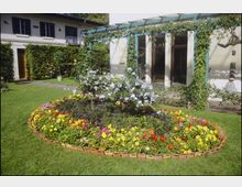 Ein gepflegter Garten mit einem runden Blumenbeet im Vordergrund, das bunt blühende Pflanzen und einen Strauch mit lila Blüten enthält. Im Hintergrund ein Gebäude mit begrüntem Glasvordach, großen Fenstern und einer Hecke, die den Garten umrahmt.