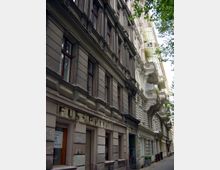 Alt-Text: Straßenansicht einer Altbaufassade mit detailreicher, historischer Architektur. Die Gebäude haben mehrere Stockwerke, hohe Fenster mit Holzrahmen und Putzverzierungen; teils sind Balkone mit Geländern sichtbar. Ein verblasstes Schild mit der Aufschrift „Fußpflege“ ist an einem der unteren Geschosse erkennbar.