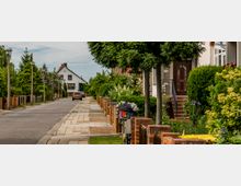 Wohnstraße in einem Vorstadtgebiet mit Einfamilienhäusern, Vorgärten und Bäumen. Die Straße ist von Zäunen und Hecken gesäumt, ein geparktes Auto und ein Fahrrad mit Blumenkorb befinden sich im Vordergrund.