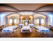 Ein heller Veranstaltungsraum mit gewölbter Decke und großen Fenstern, die durch moderne Glasrahmen unterteilt sind. In der Mitte stehen mehrere gedeckte Tische mit weißen Tischdecken und blauen Stühlen auf einem gemusterten Holzfußboden.