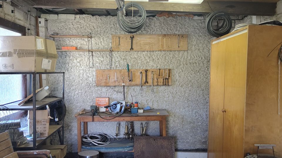 Innenansicht einer kleinen Werkstatt oder Abstellkammer mit freiliegenden Holzbalken und Deckenleuchten, einer Werkbank mit Werkzeugleisten an einer rau verputzten Wand sowie aufgehängten Kabelrollen. Links stehen Regale mit Kartons, rechts ein hoher Holzschrank und ein runder Grill; am Boden liegen Geräte und diverse Gegenstände.