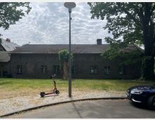 Eine alte, einstöckige Backsteinhalle mit kleinen vergitterten Fenstern, umgeben von einer teilweise bewachsenen Grünfläche. Im Vordergrund stehen ein E-Scooter an einer Straßenlaterne und ein geparktes Auto am Straßenrand.