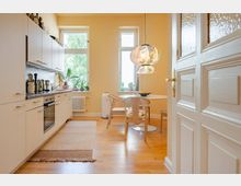 Helle Wohnküche mit weißen Einbauschränken entlang einer Wand, Holzfußboden und kleinem Essbereich mit rundem Tisch und Stühlen. Zwei hohe Fenster bringen Tageslicht hinein; über dem Tisch hängen gemusterte Papierlampen, daneben sind Pflanzen und eine weiße Tür im Vordergrund zu sehen.