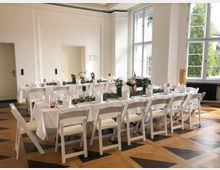 Heller, hoher Festsaal mit weißen Wandpaneelen und großen Sprossenfenstern, durch die Tageslicht und Blick ins Grüne fällt. Zwei lange Tafeln mit weißen Tischdecken, gefalteten Servietten, Blumen- und Kerzendekoration stehen auf einem gemusterten Holzparkettboden.